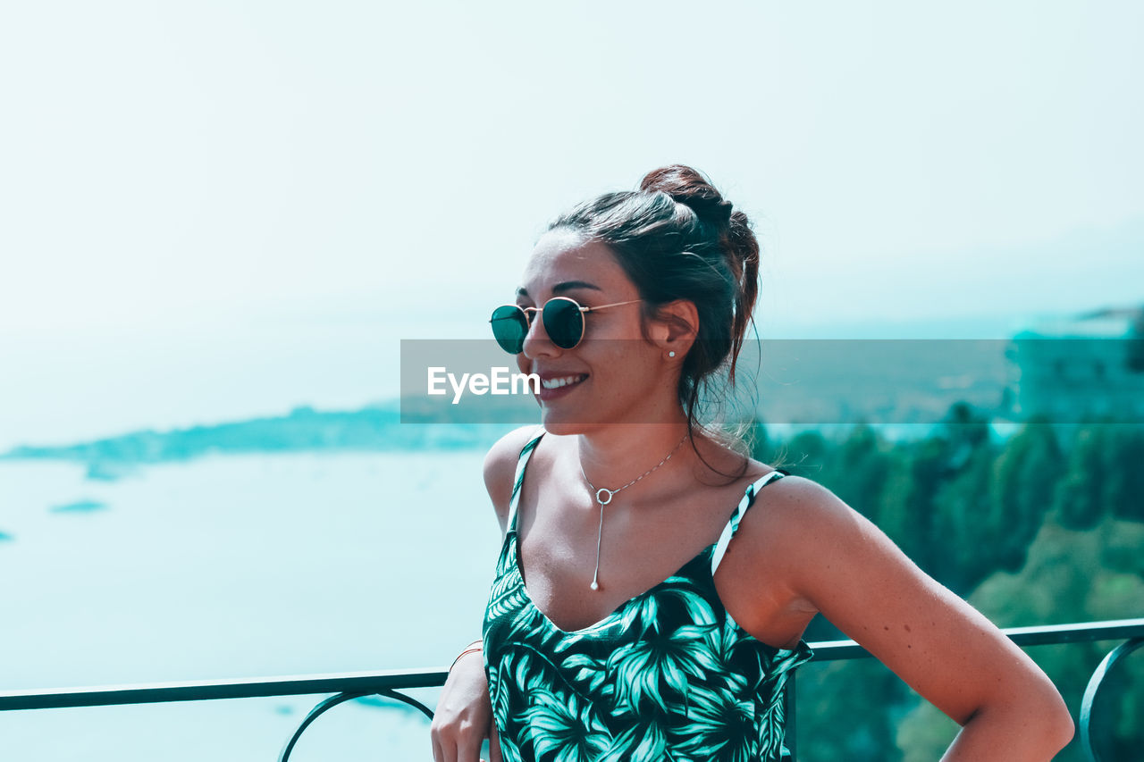 Smiling woman looking away while standing by railing against sky