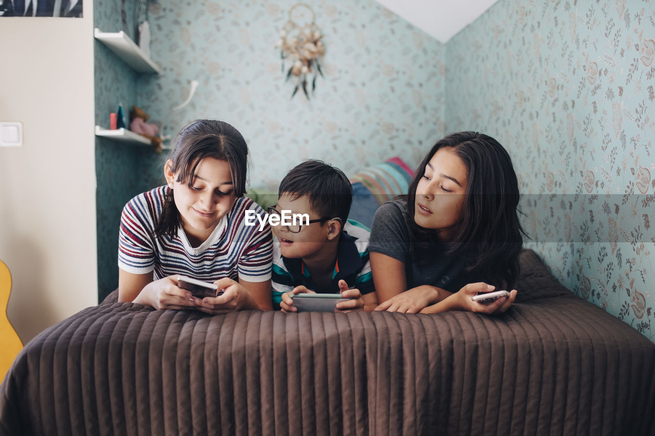 Siblings using wireless technologies while lying on bed by wall at home