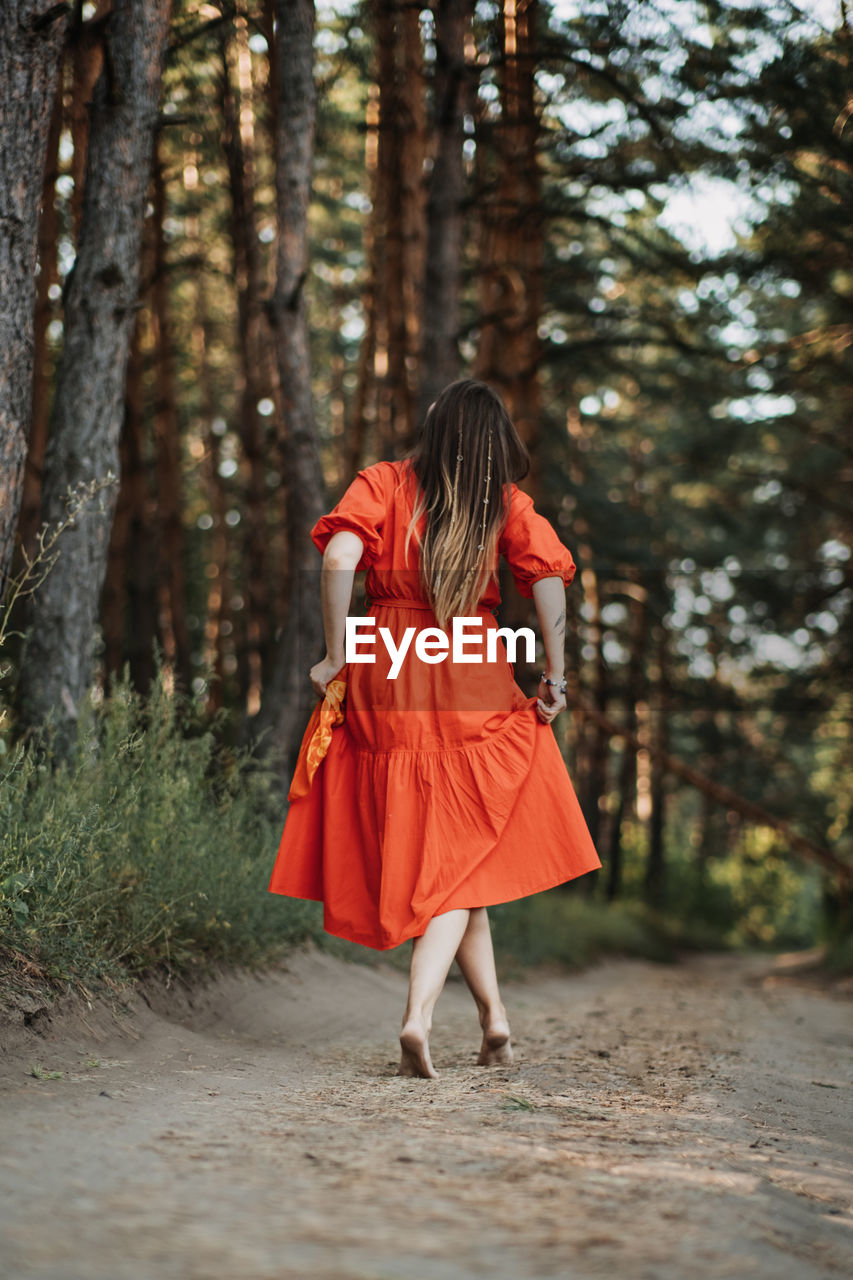 Barefoot happy young woman in red dress with hand raised dancing in pine forest at summer day.