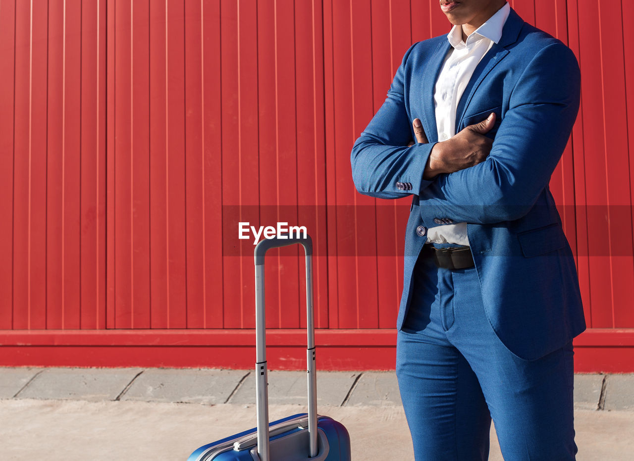 Crop unrecognizable black businessman in classy blue suit standing with arms crossed near suitcase against red wall