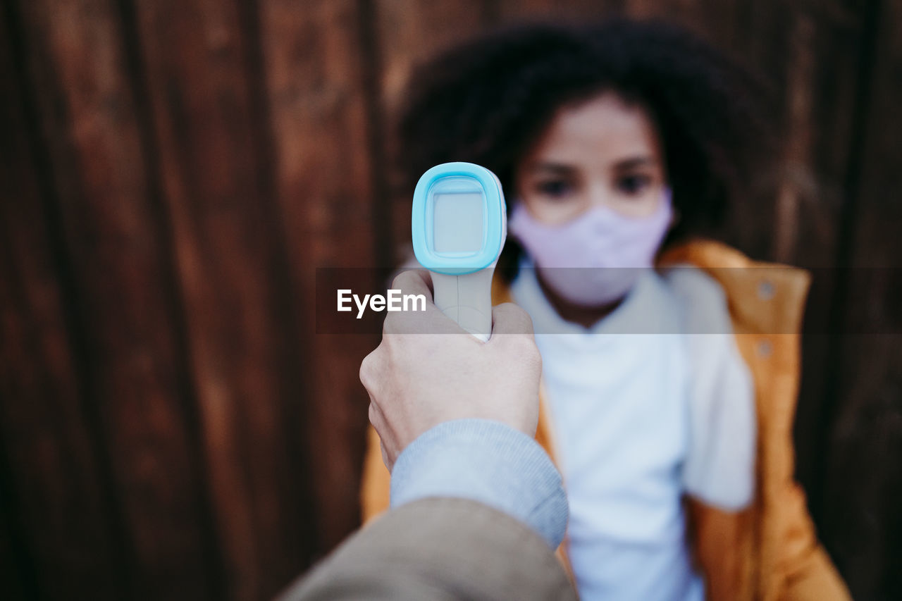 Woman using infrared thermometer while checking girl temperature against wooden wall