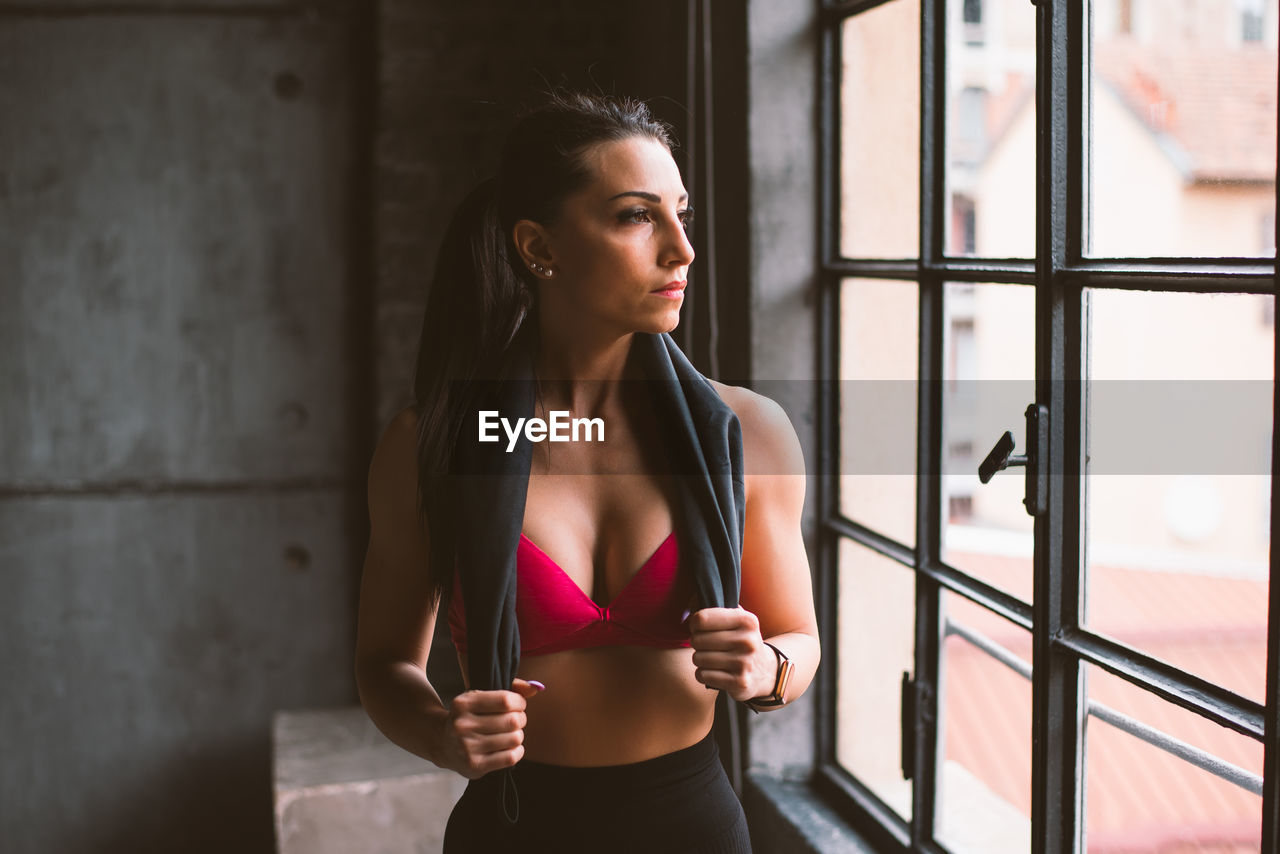 BEAUTIFUL YOUNG WOMAN LOOKING AWAY WHILE STANDING AGAINST WINDOW