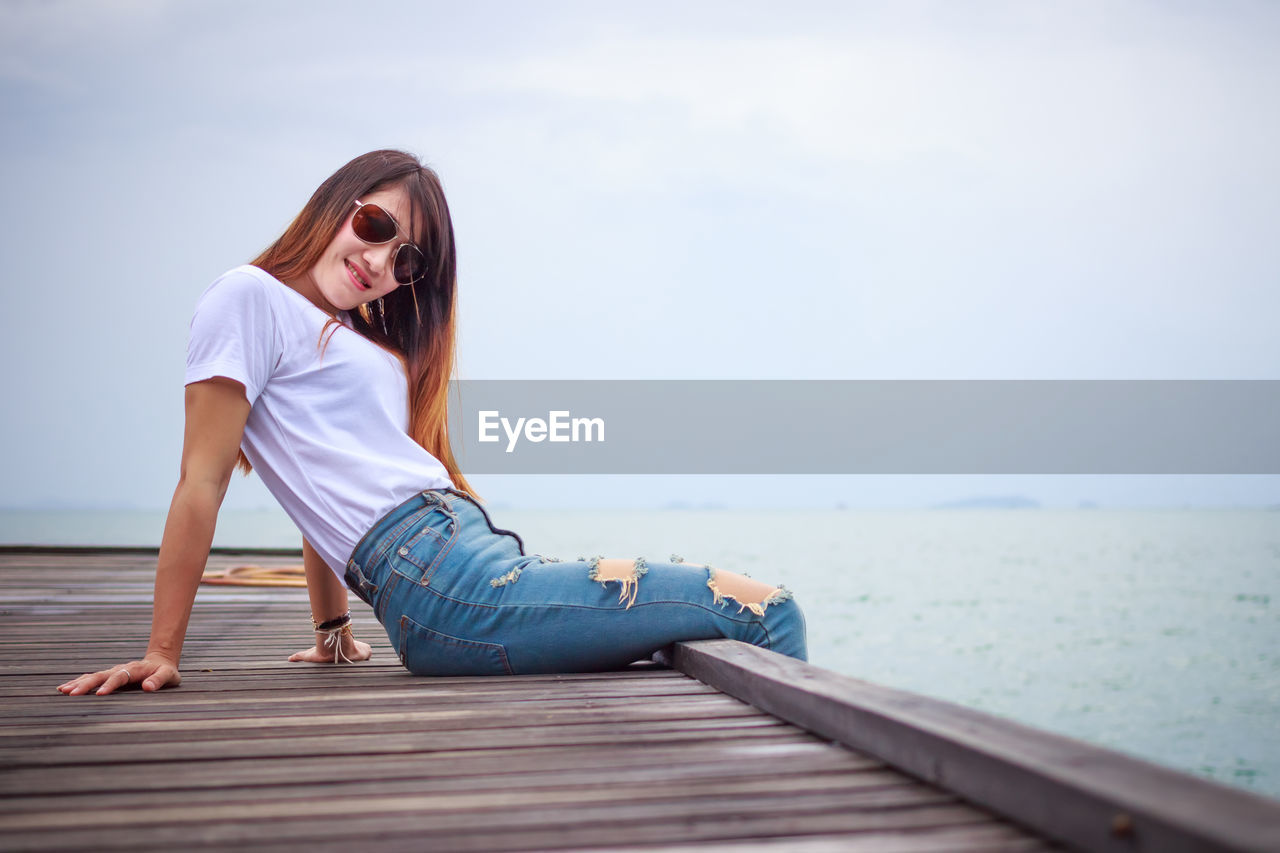 Young woman wearing sunglasses sitting on pier by sea against sky