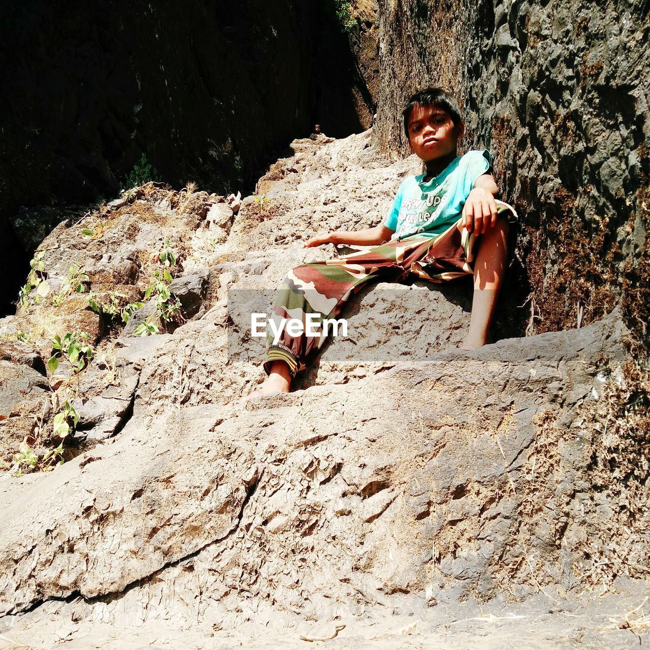Low angle portrait of boy sitting on rock formation during sunny day