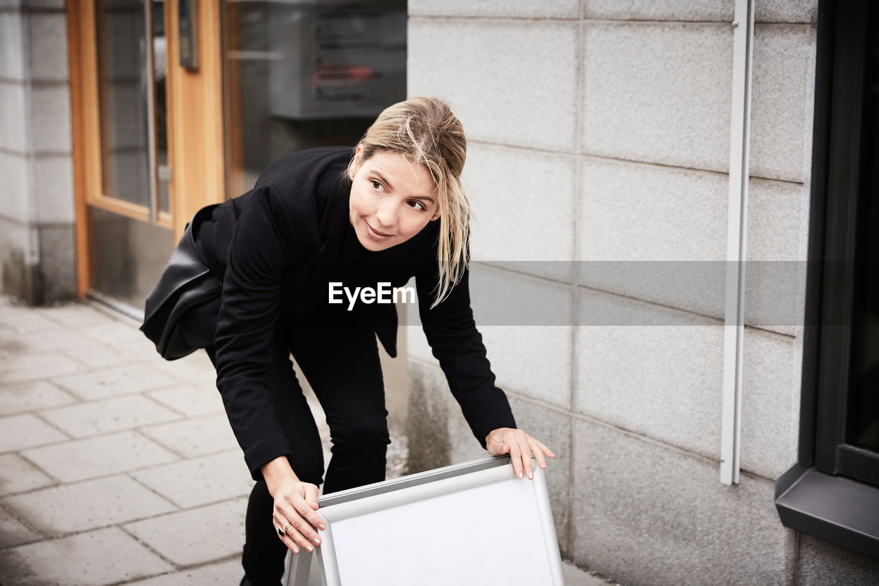Female real estate agent looking away while arranging blank signboard on sidewalk