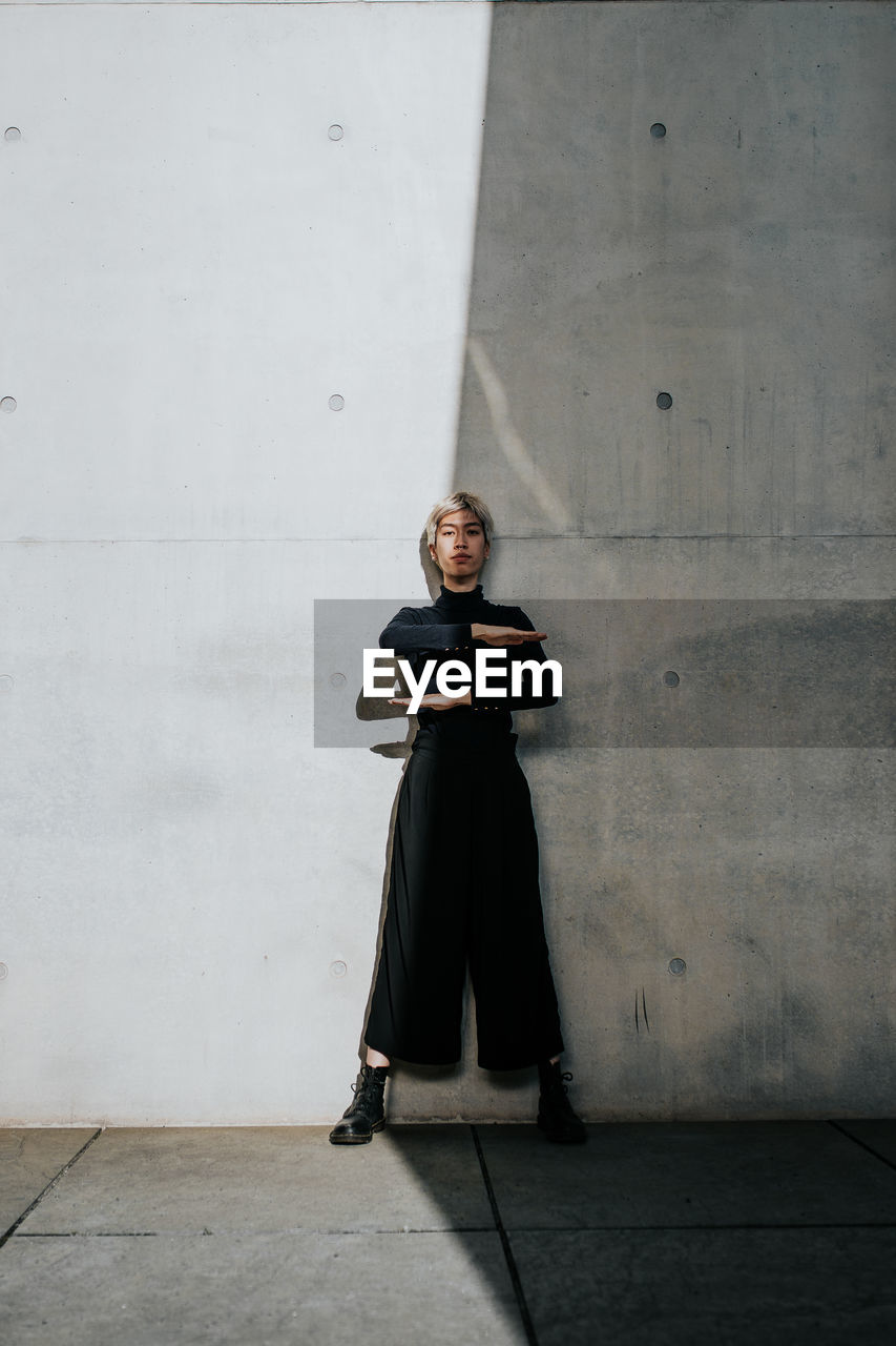 Portrait of young woman gesturing equal sign while standing against wall