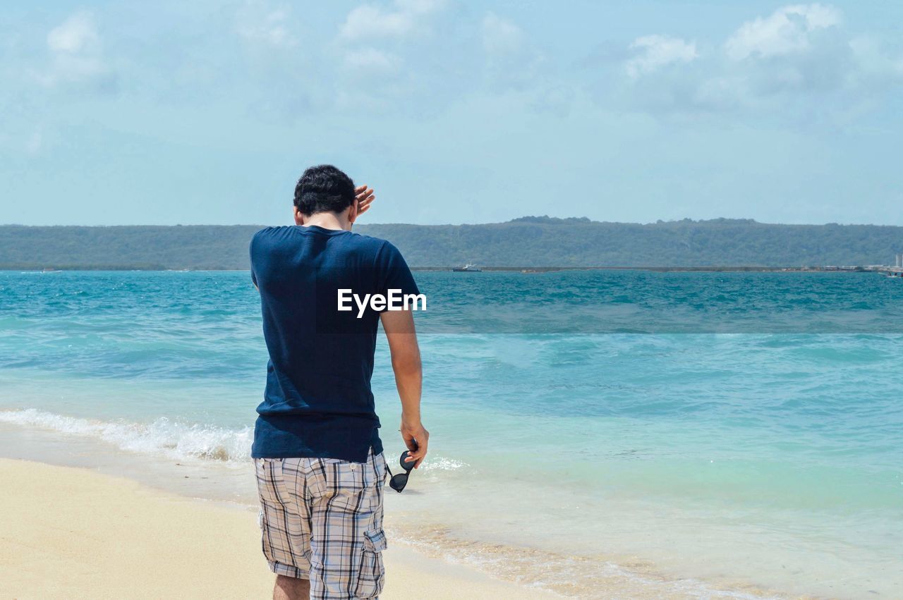 Rear view of man standing at sea shore against blue sky