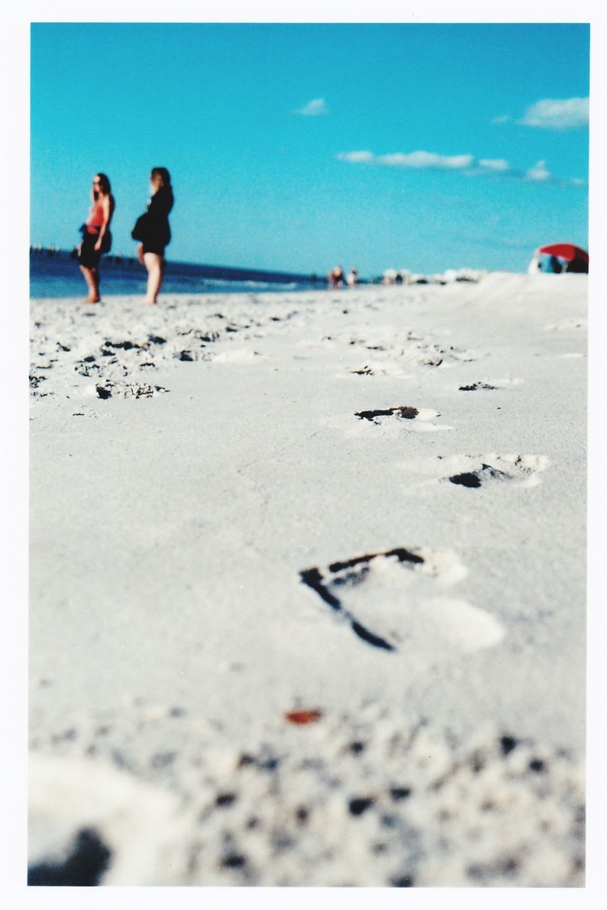 Footprint at beach leading towards women against sky on sunny day