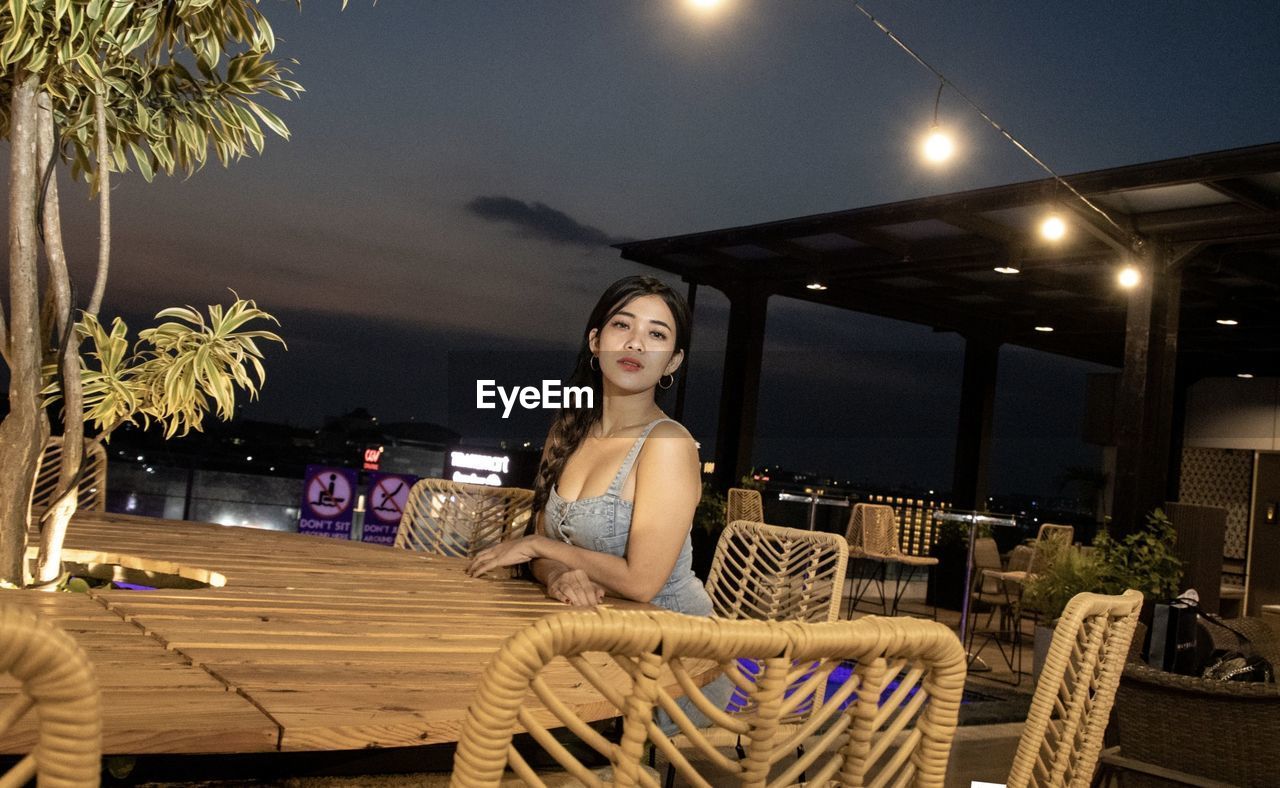one person, adult, night, young adult, women, furniture, seat, sitting, smiling, portrait, illuminated, chair, table, looking at camera, happiness, lifestyles, three quarter length, food and drink, relaxation, nature, fashion, lighting equipment, architecture, clothing, leisure activity, emotion, business, city