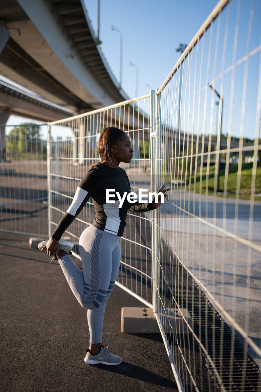 Black sportswoman stretching near fence