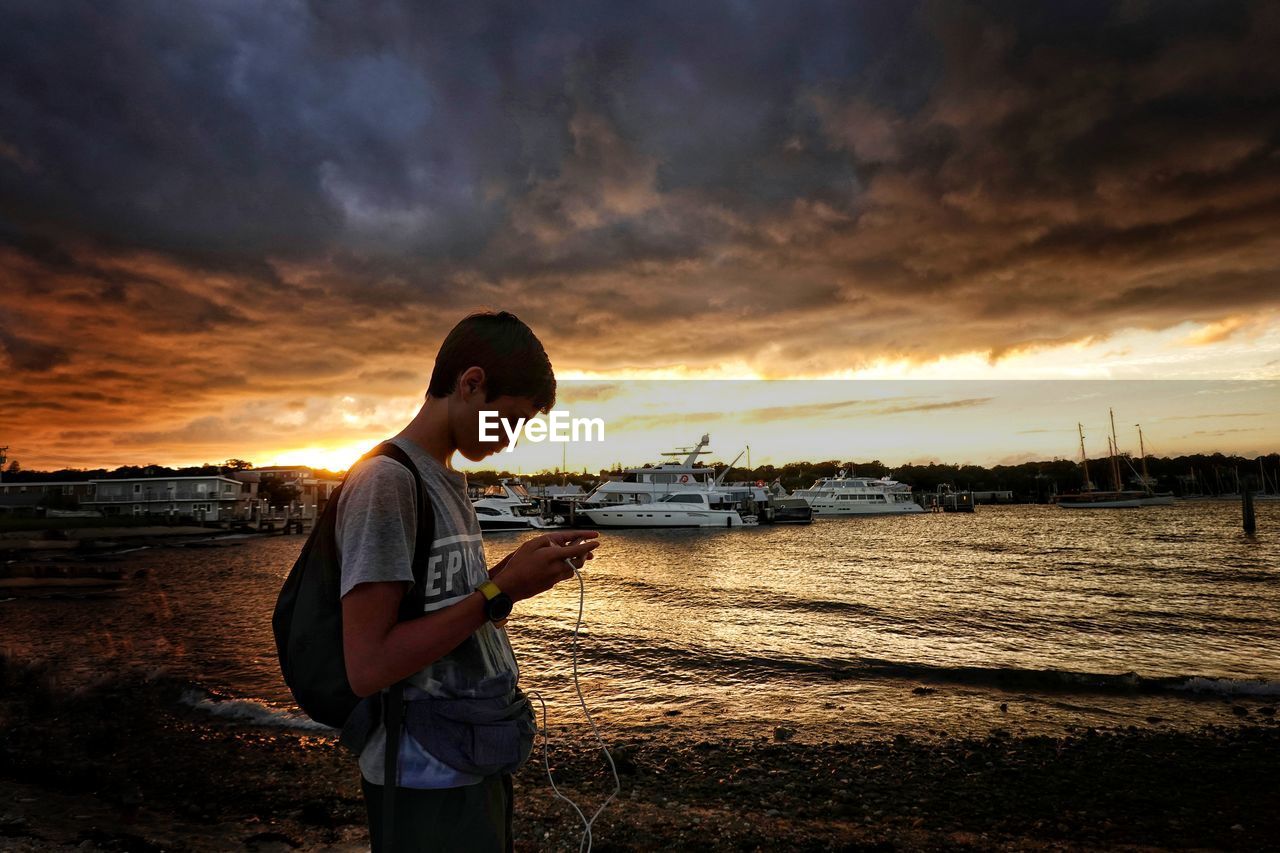 Side view of boy standing at beach against cloudy sky during sunset