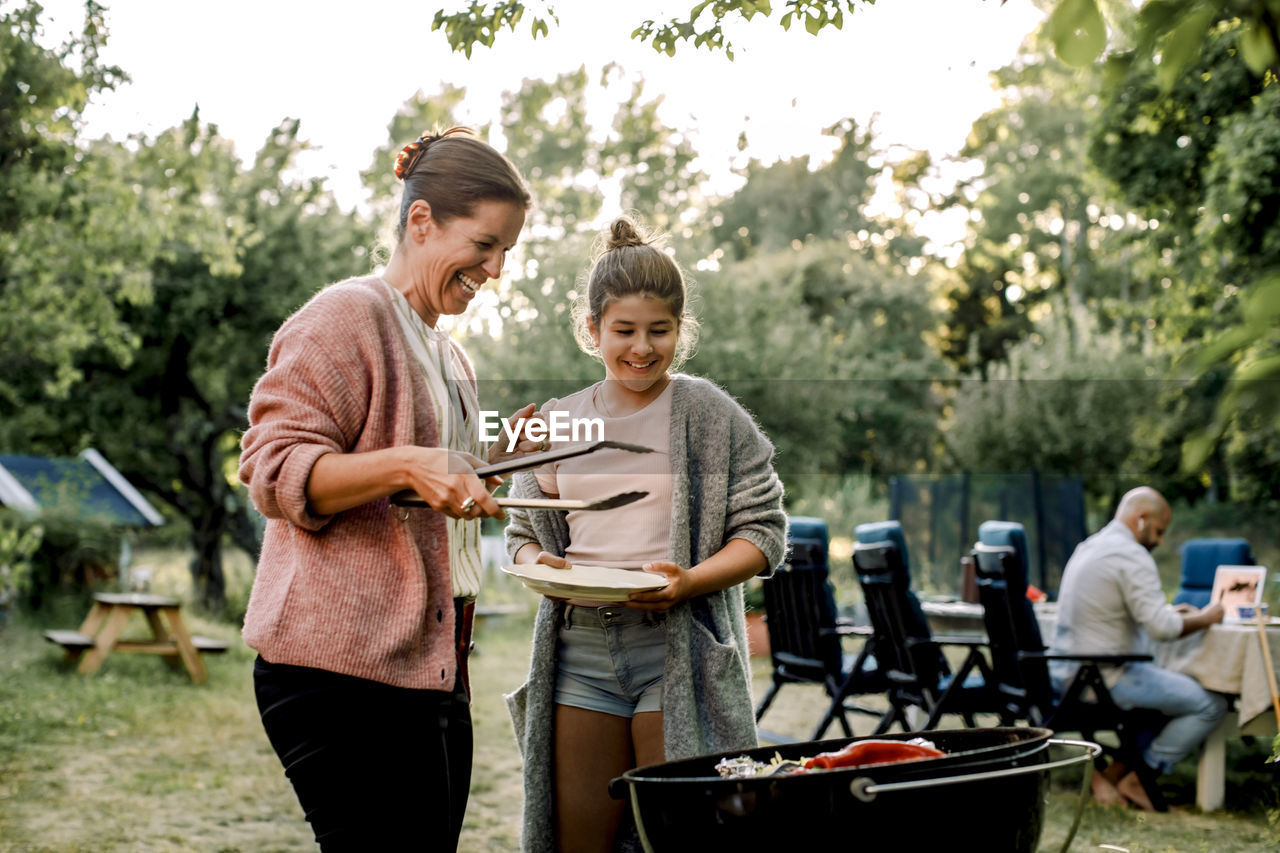 Smiling mother and daughter looking at red chili pepper over barbecue grill in yard