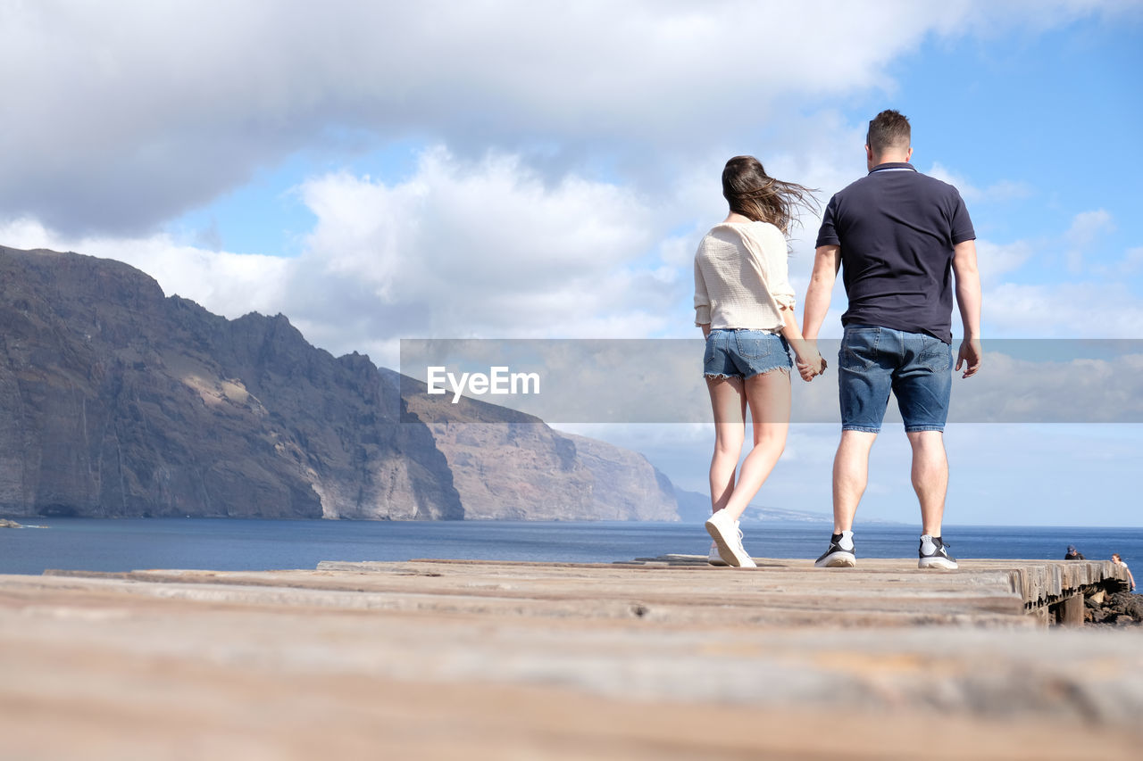 REAR VIEW OF COUPLE WALKING ON MOUNTAIN BY SEA