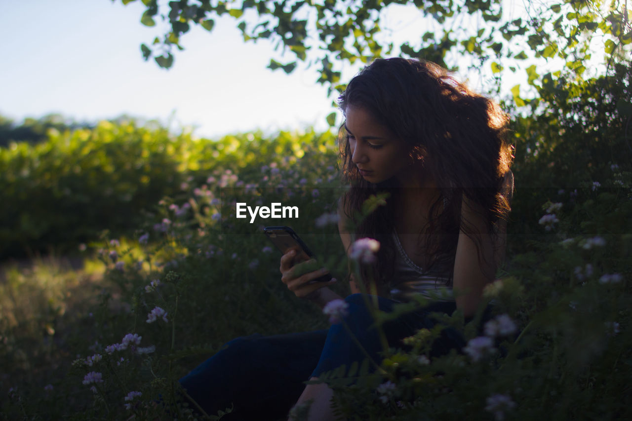 Woman using phone while sitting on field by flower