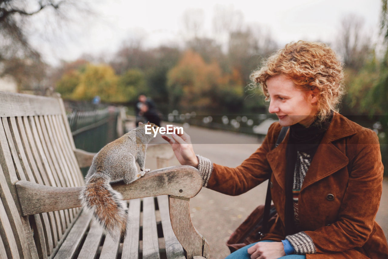 Smiling woman playing with squirrel on bench in park