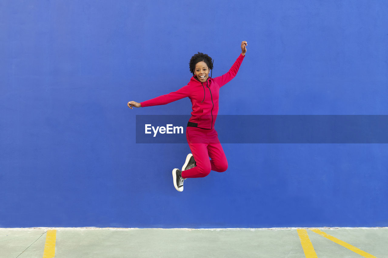 Happy woman jumping on footpath in front of blue wall