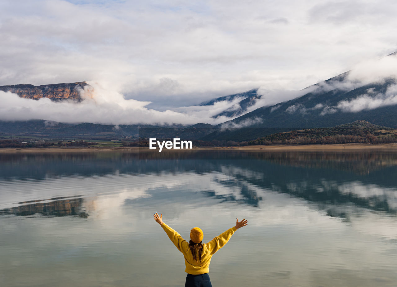 Rear view of unrecognizable woman with arms outstretched up looking at mountains reflected in lake