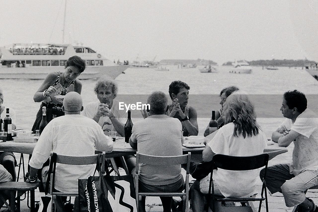 REAR VIEW OF PEOPLE SITTING ON CHAIR AT SEA