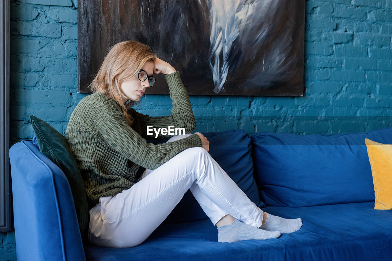 Young thoughtful caucasian woman in green sweater and white jeans sitting on sofa with eyes closed 