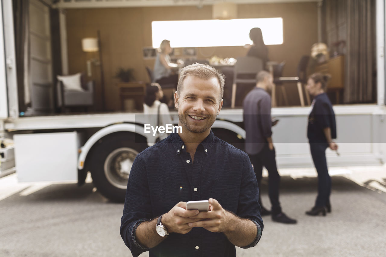 Portrait of smiling businessman holding mobile phone with colleagues and portable office truck on road in background