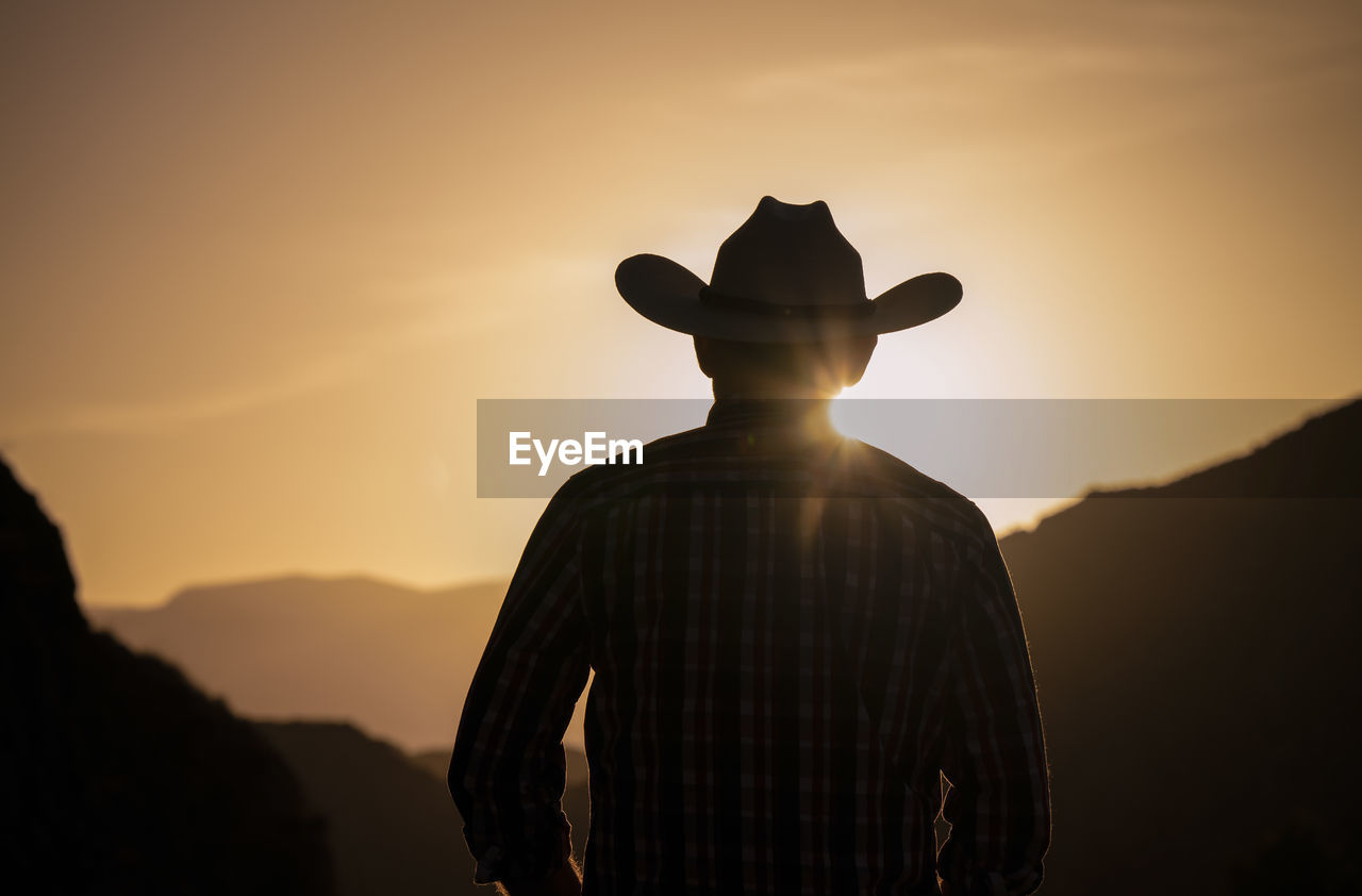 Silhouette of adult man in cowboy hat against mountain and sky during sunset