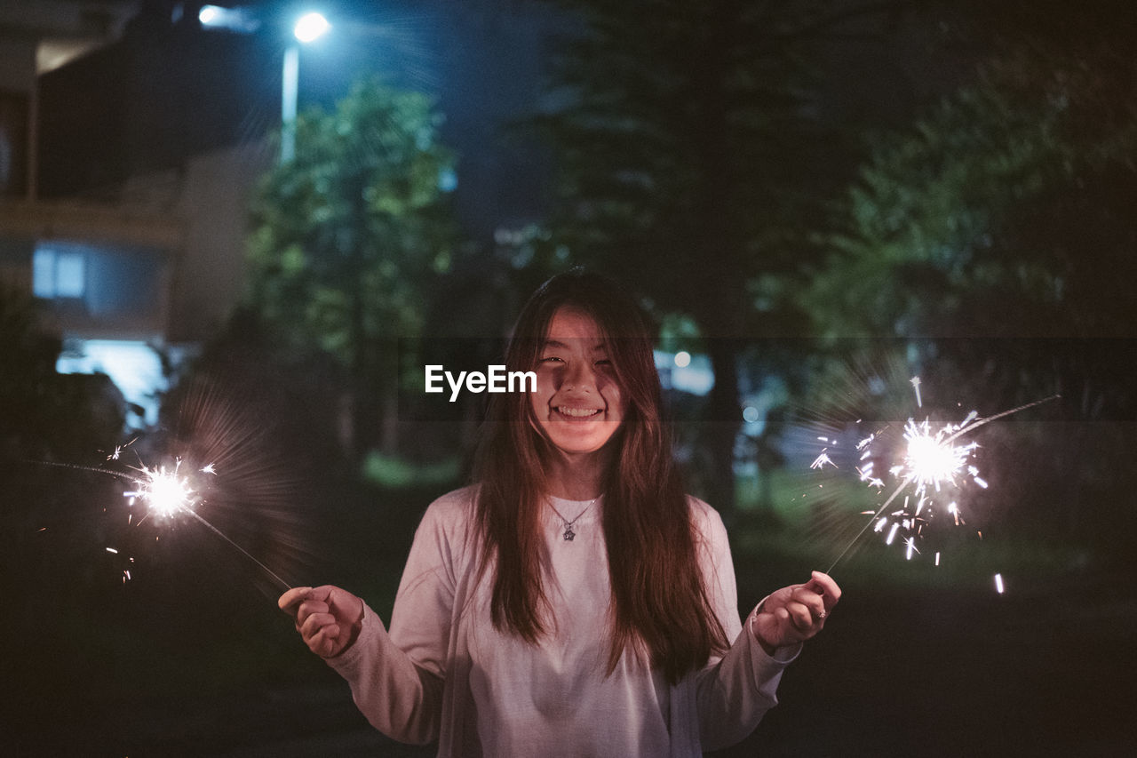 Young woman holding illuminated sparkler while standing at night