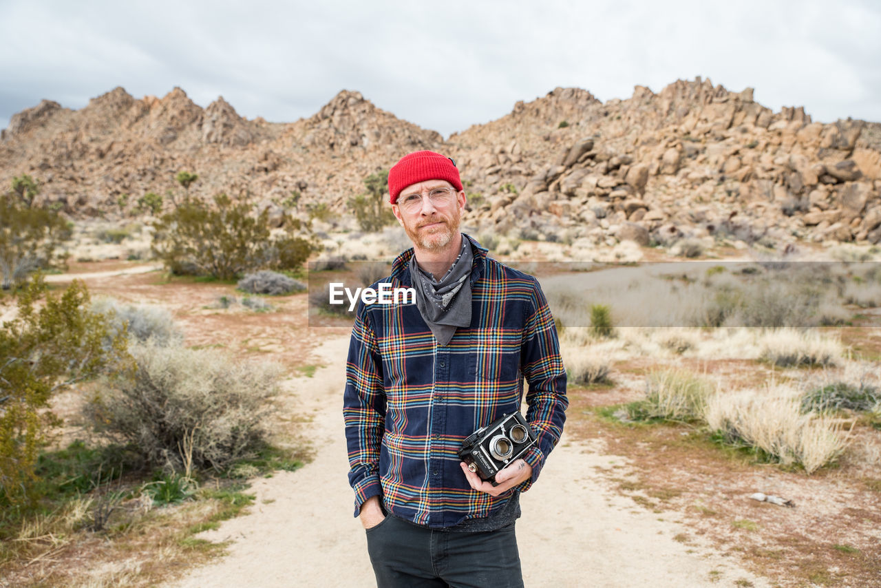 Man in beanie with beard and glasses by far off stone piles in desert