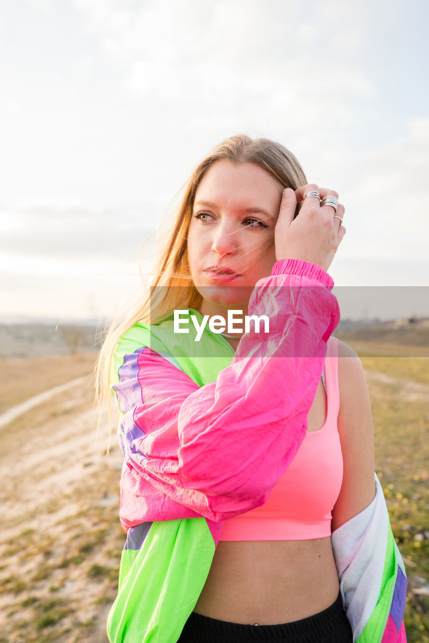 Contemporary blond haired young woman in pink bra with colorful jacket looking away with remote land on background