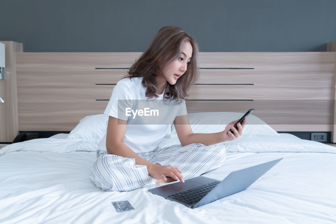 YOUNG WOMAN USING MOBILE PHONE ON BED AT HOME