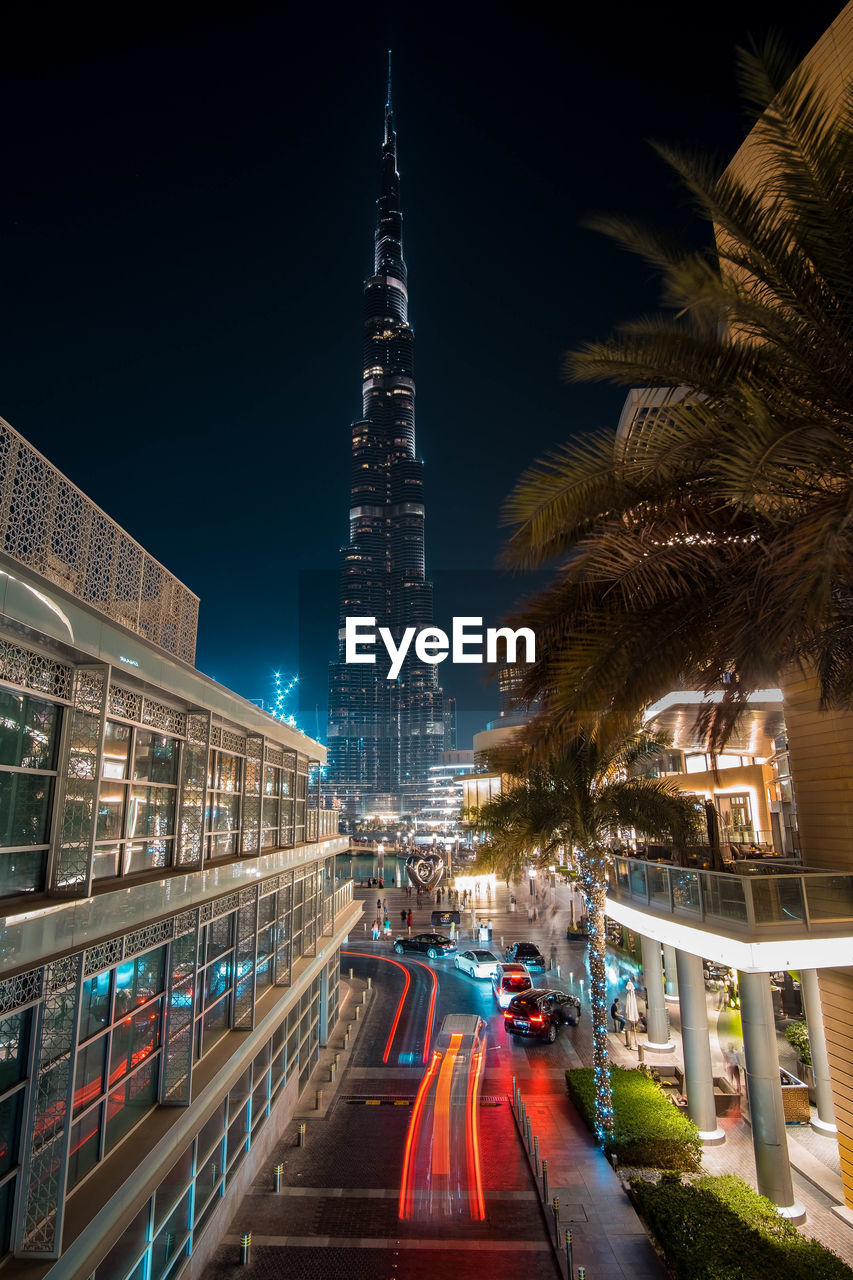 LIGHT TRAILS ON ROAD AMIDST BUILDINGS AGAINST SKY