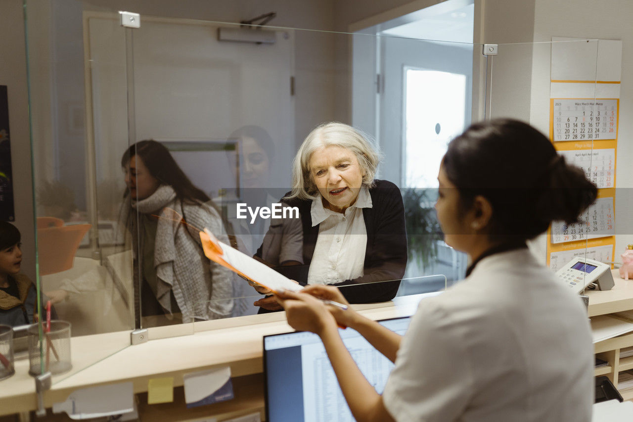 Senior female patient understanding form filling procedure by receptionist through transparent shield in clinic