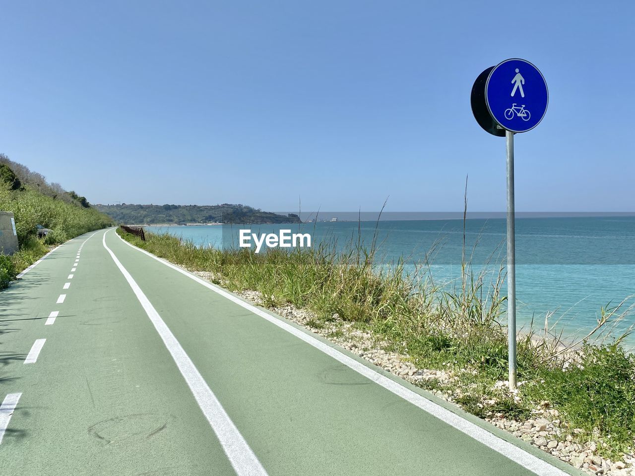 road, sky, sign, road sign, symbol, water, nature, sea, transportation, blue, clear sky, land, beach, no people, day, coast, guidance, scenics - nature, tranquility, horizon, the way forward, tranquil scene, beauty in nature, sunny, horizon over water, communication, plant, road marking, outdoors, travel, marking, traffic sign, arrow symbol, sunlight, vacation, grass, travel destinations, directional sign, walkway, highway, copy space, shore, infrastructure, non-urban scene, environment