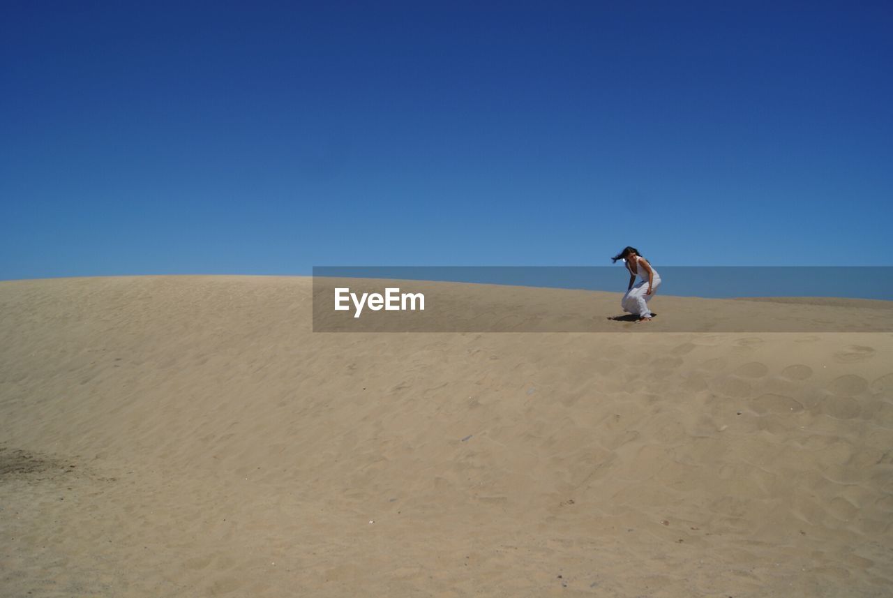Low angle view of young woman sandboarding against clear blue sky at desert