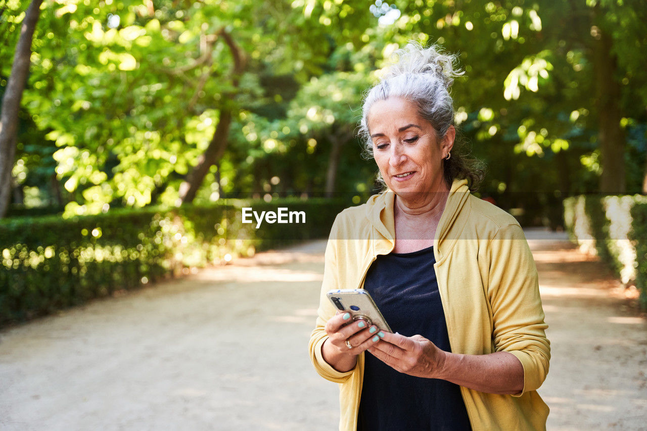 Positive elderly female athlete with gray hair smiling and reading message in social media on smartphone while standing on park alley during break in fitness training