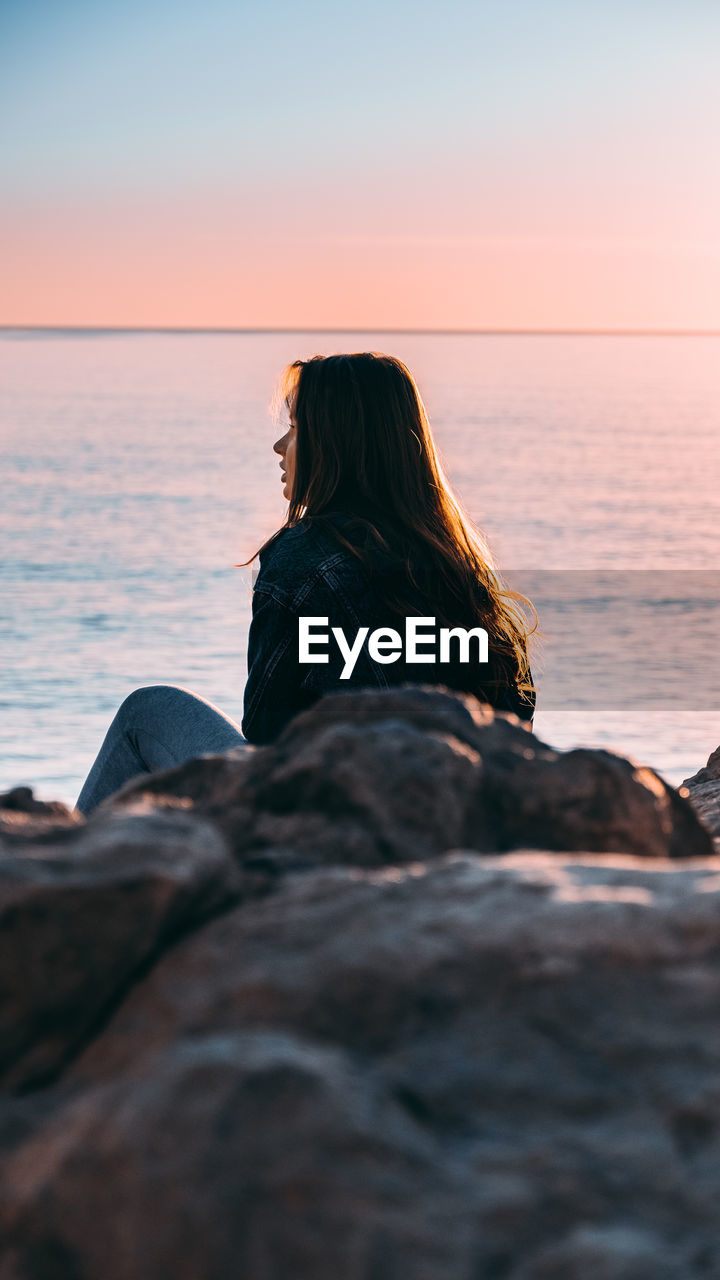 Midsection of woman sitting by sea against sky during sunset