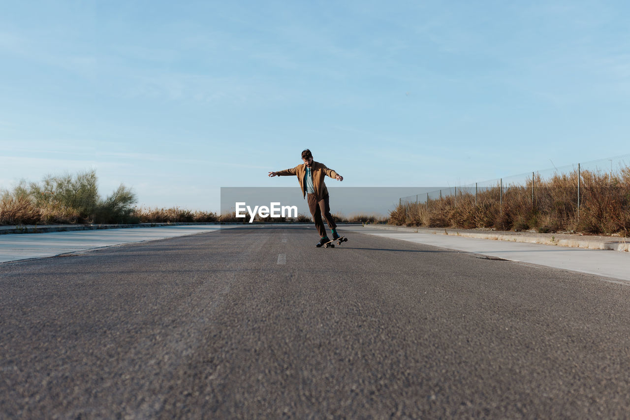 Full body young bearded male skater in stylish wear riding skateboard along asphalt road in countryside