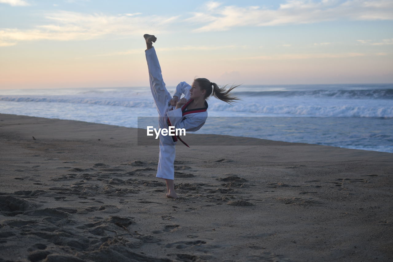 Woman practicing karate on beach against sky during sunset