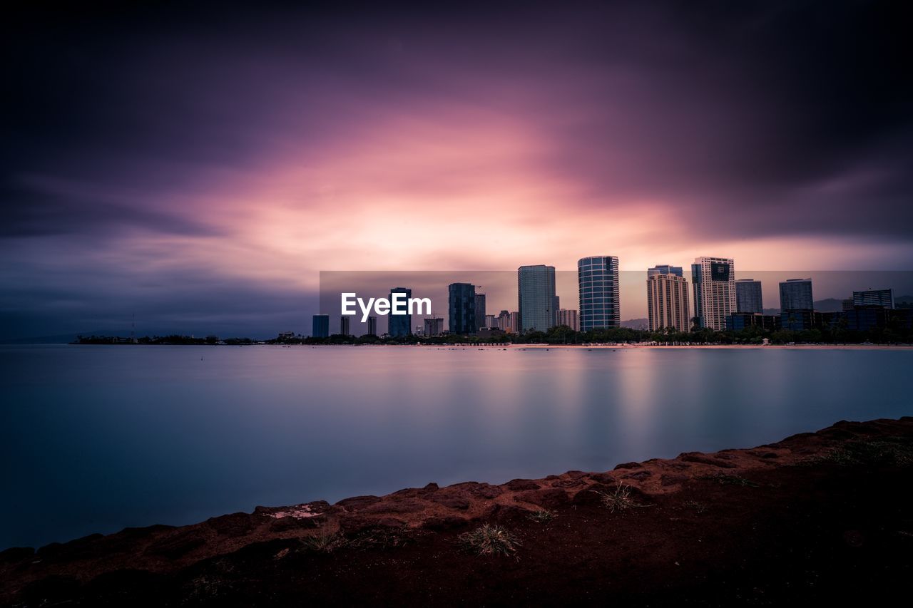 Sea and buildings against cloudy sky during sunset