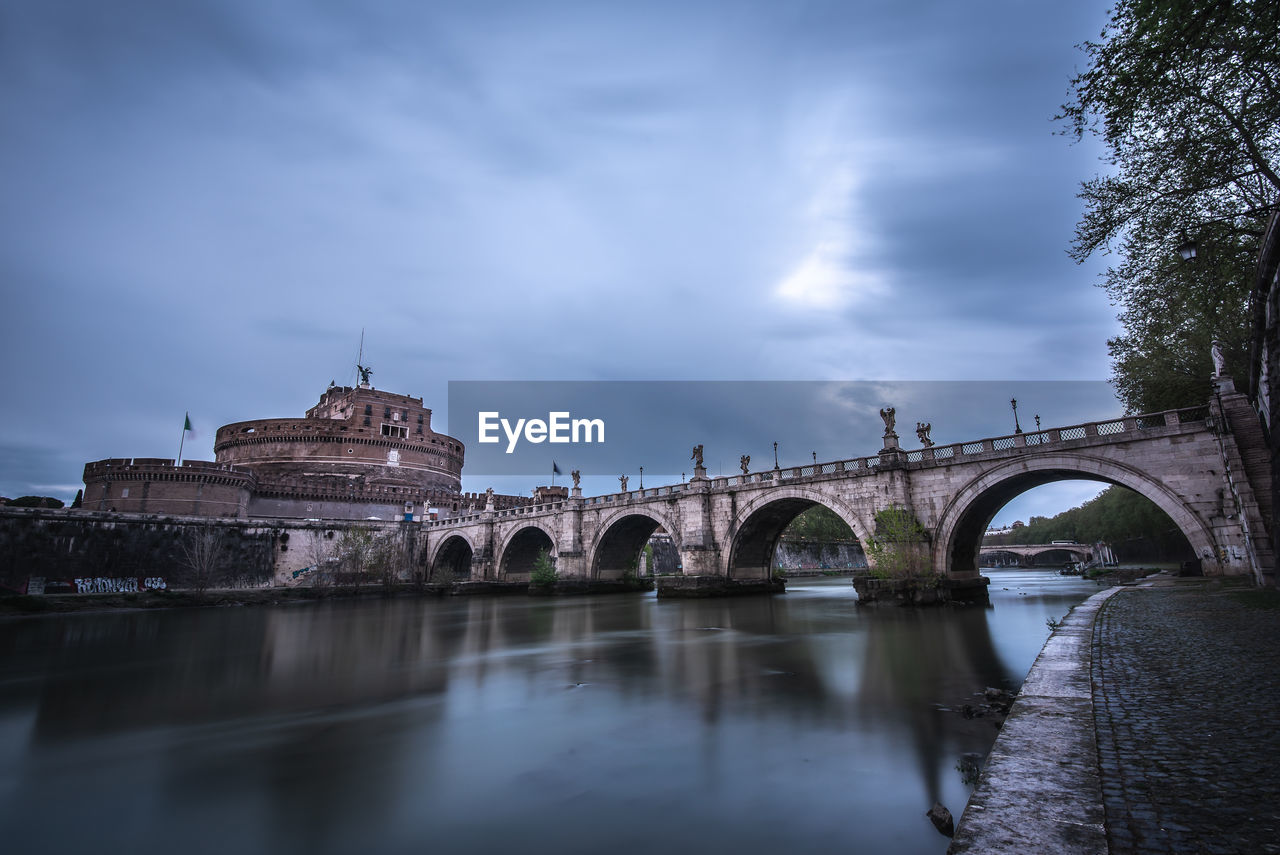 Arch bridge over river by buildings in city during sunset