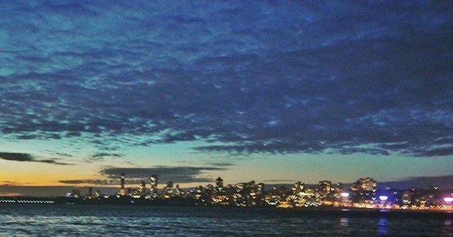 VIEW OF ILLUMINATED CITYSCAPE AGAINST CLOUDY SKY AT DUSK