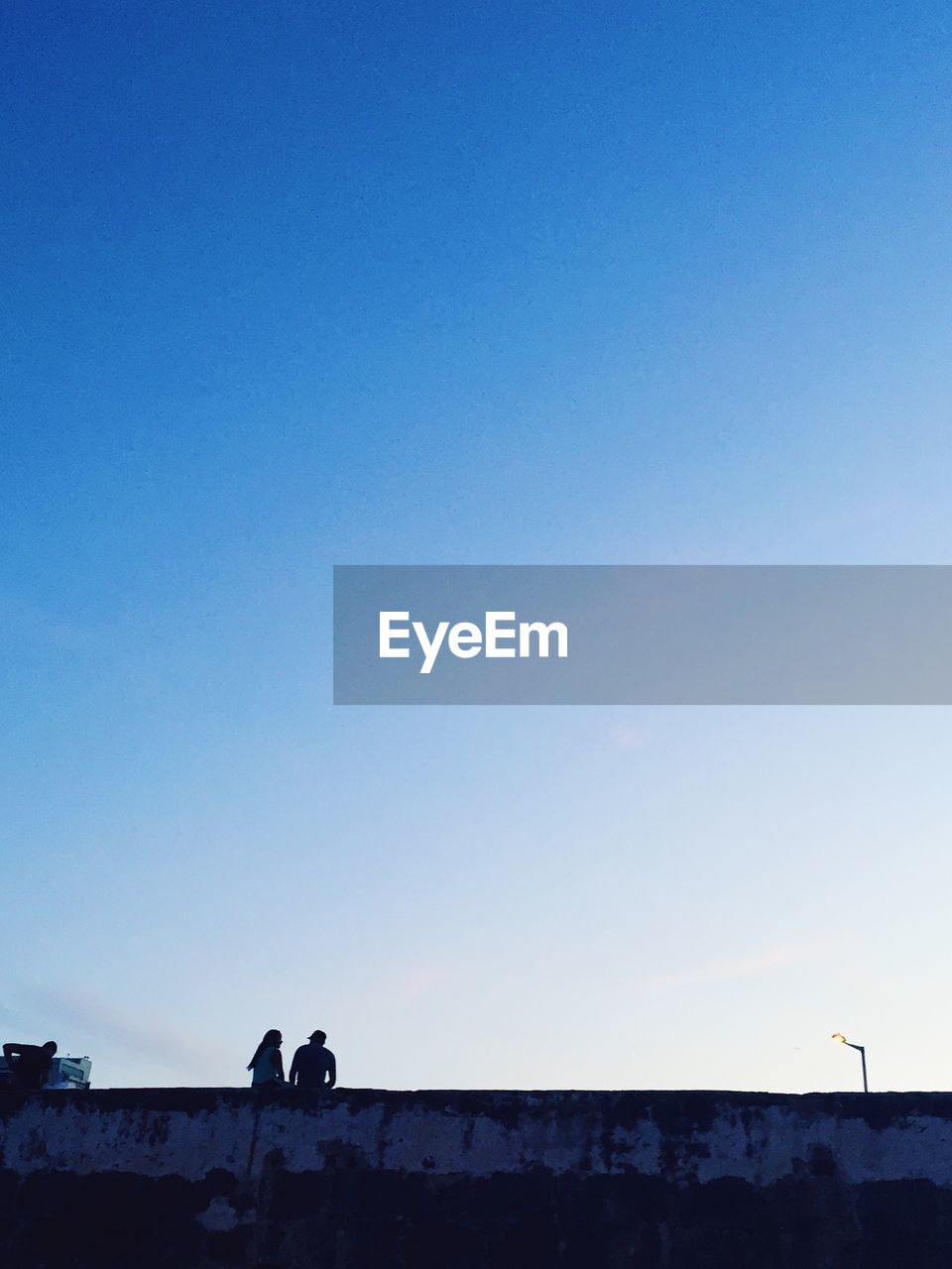 Rear view of man and woman sitting on retaining wall against sky