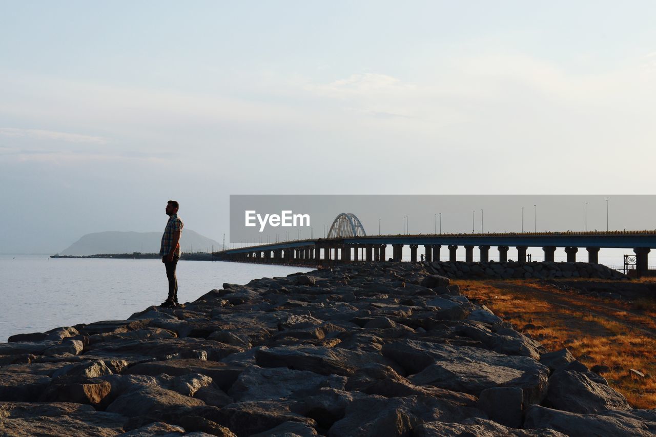 Man standing on rock by bridge against sky