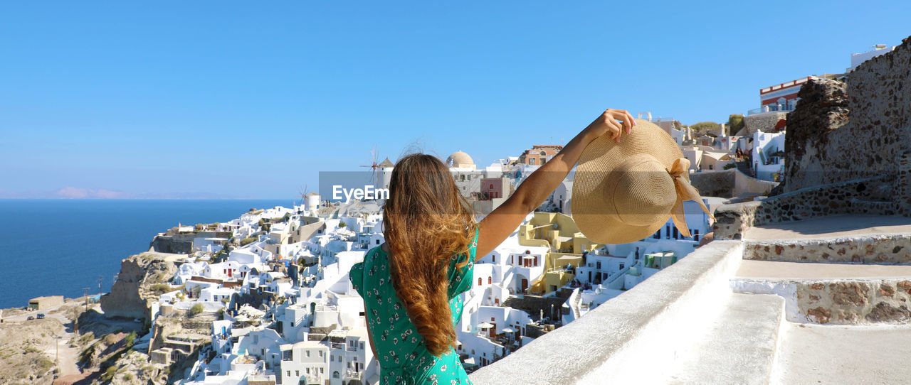Rear view of woman holding hat while looking at buildings by sea against sky