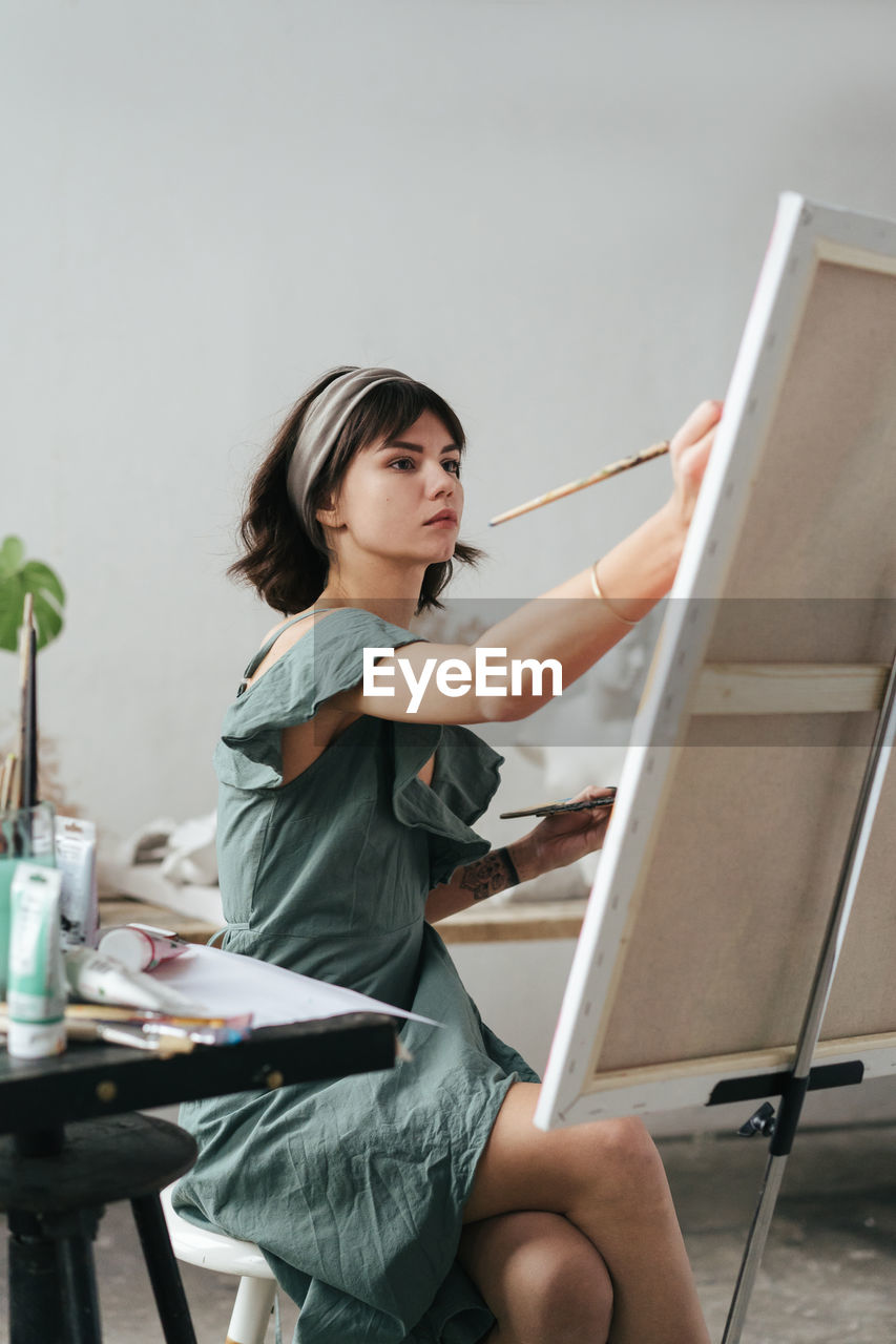 Young woman painting on canvas while sitting on table