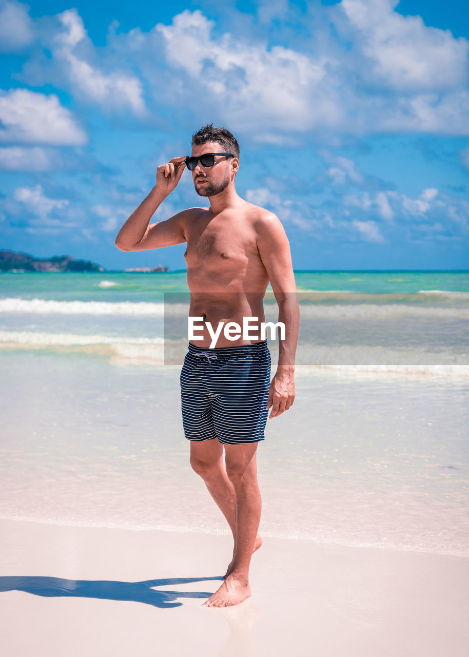 Full length of shirtless man in sunglasses walking at beach against sky