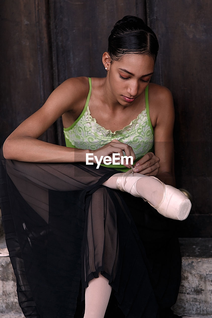 Young woman wearing ballet shoes while sitting against wall