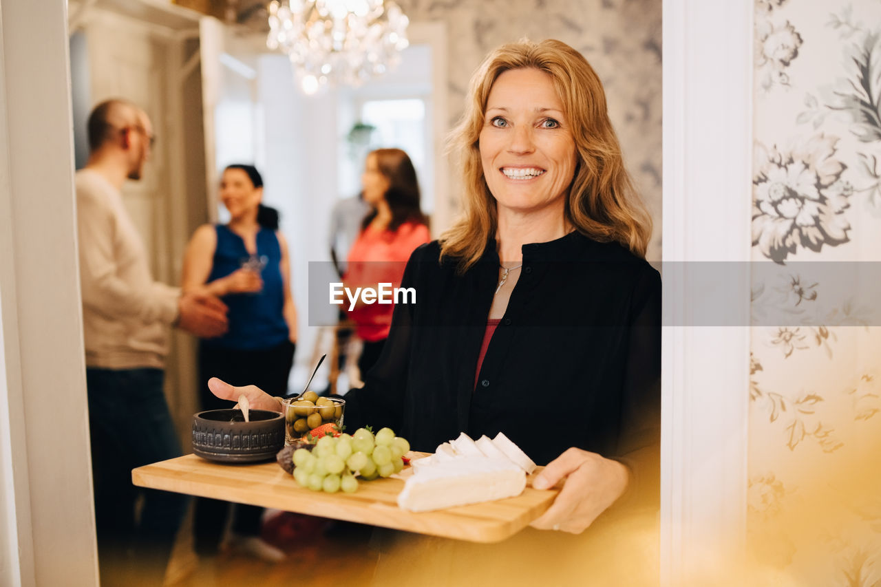 Portrait of smiling mature woman holding serving tray with friends standing in background