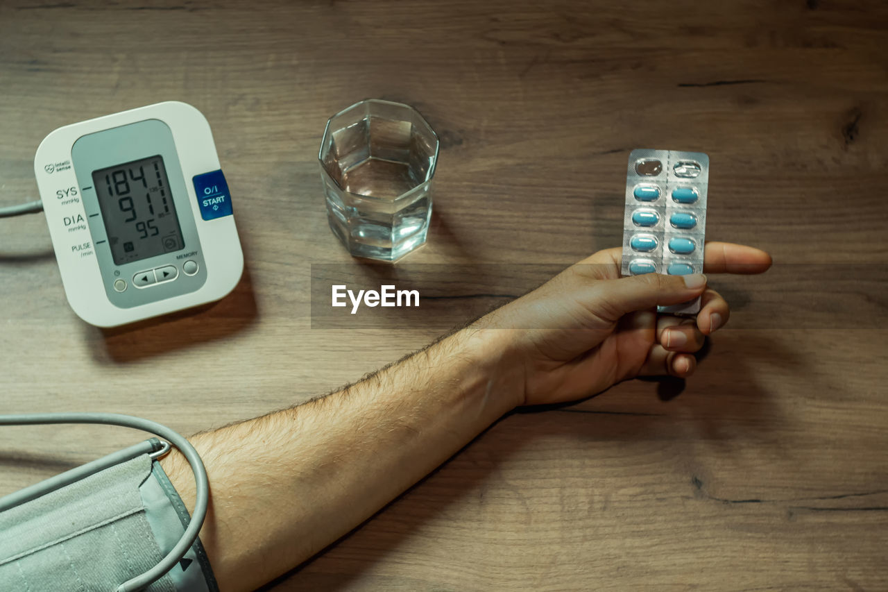 A man's hand with a pill in his hand, a blood pressure monitor and a glass of water on a wooden