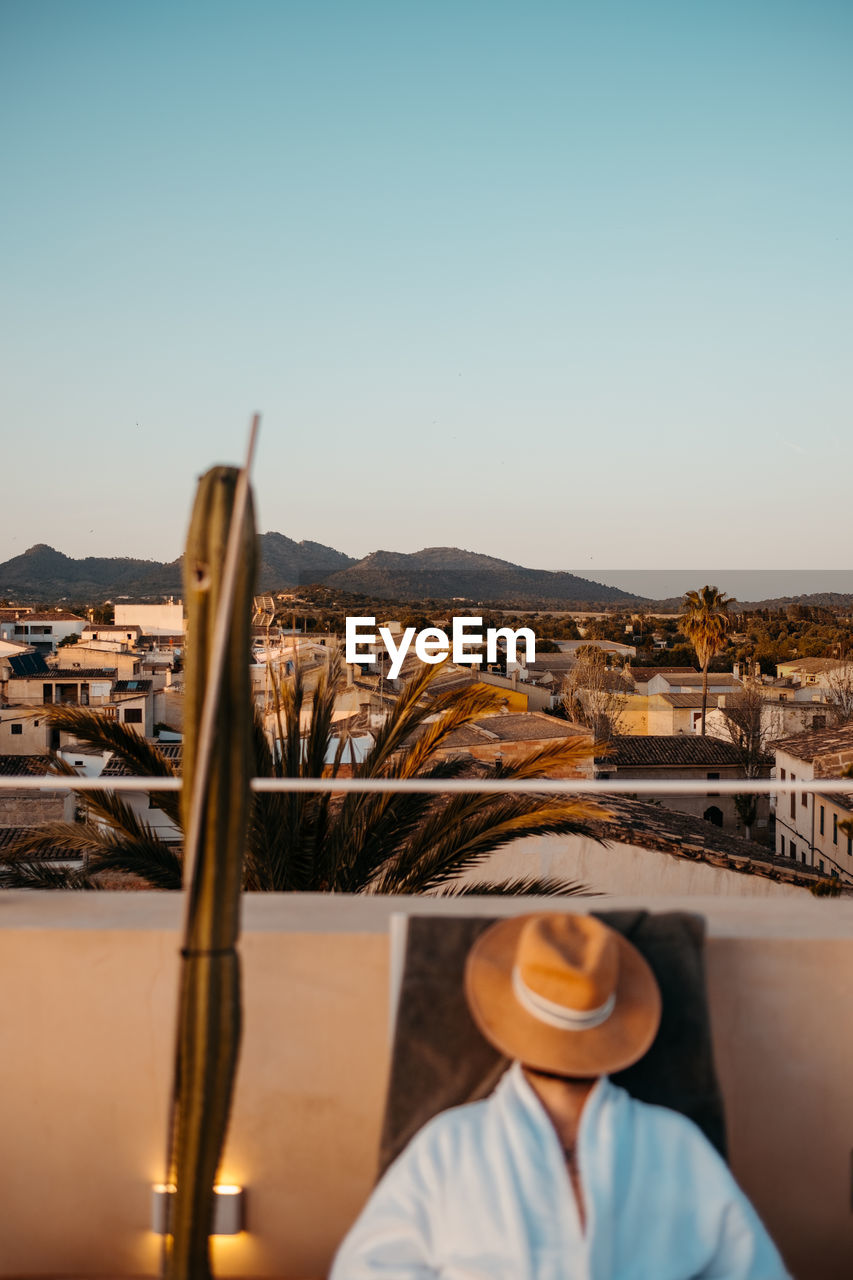 Anonymous traveler in robe with straw hat on face lying on sunbed while relaxing on rooftop against cloudless sunset sky during holidays in mallorca person