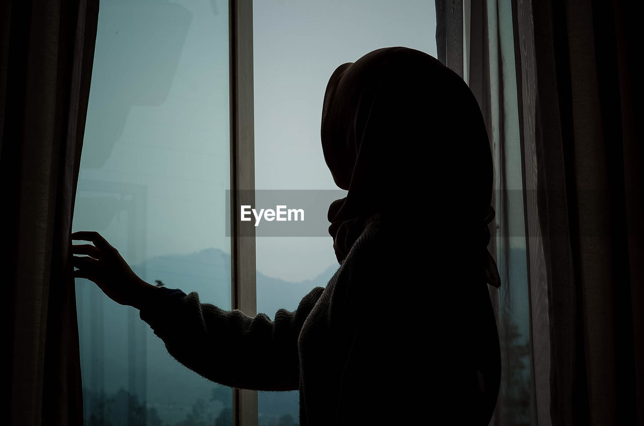 Portrait of silhouette woman looking through window at hotel